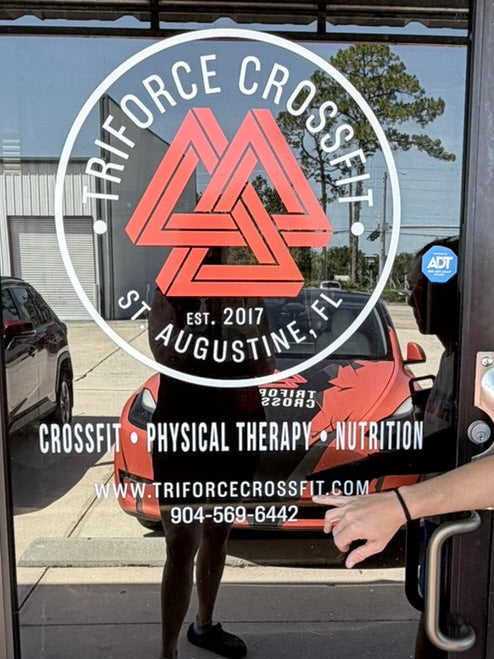 A person in athletic wear is opening a glass door at a fitness center. The door has a large circular logo for Triforce Crossfit in St.