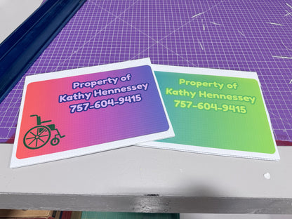 Two printed cards with colorful gradient backgrounds and the text 'Property of Kathy Hennessey 757-604-9415' are shown on a tabletop. One card has a red-to-purple-to-blue gradient with a wheelchair icon, and the other has a green-to-blue gradient.
