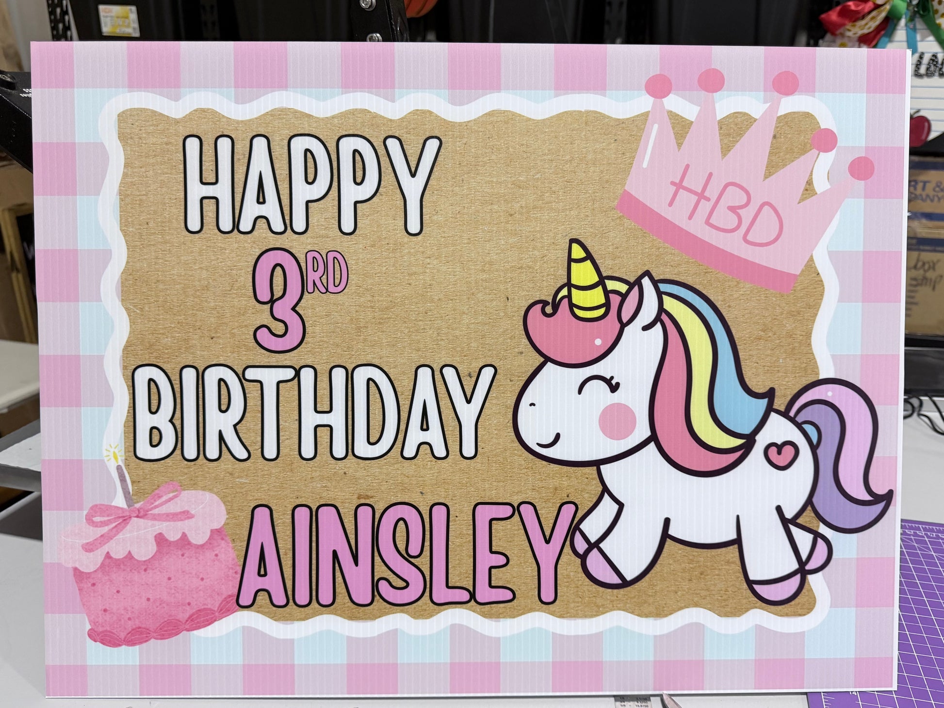 A custom printed birthday card featuring a pink and white gingham border, a brown kraft-style center panel, and a cartoon unicorn with a rainbow mane. The text reads 'HAPPY 3RD BIRTHDAY AINSLEY' with a pink crown and cupcake illustration.