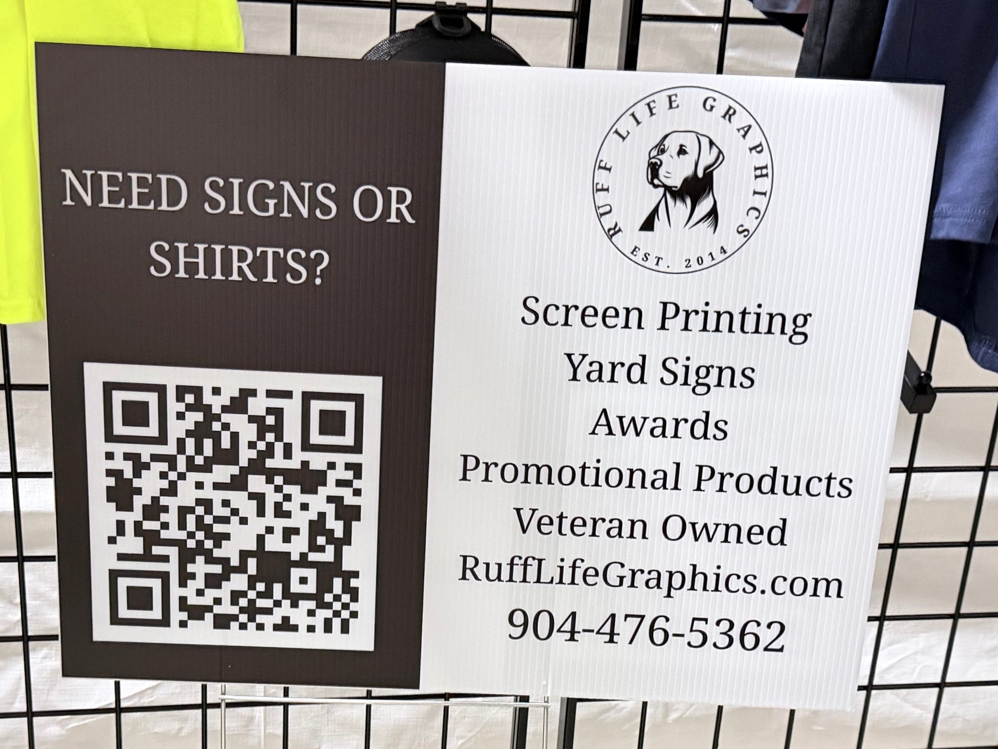 A freestanding sign on a black metal clothing rack in a store. The sign is split into two sections: the left is dark brown with the text 'NEED SIGNS OR SHIRTS?' and a QR code, and the right is white with the logo and text for 'Ruff Life Graphics' including 'Screen Printing Yard Signs Awards Promotional Products Veteran Owned' and a phone number.