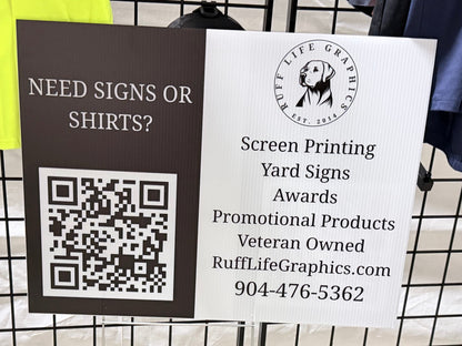 A freestanding sign on a black metal clothing rack in a store. The sign is split into two sections: the left is dark brown with the text 'NEED SIGNS OR SHIRTS?' and a QR code, and the right is white with the logo and text for 'Ruff Life Graphics' including 'Screen Printing Yard Signs Awards Promotional Products Veteran Owned' and a phone number.